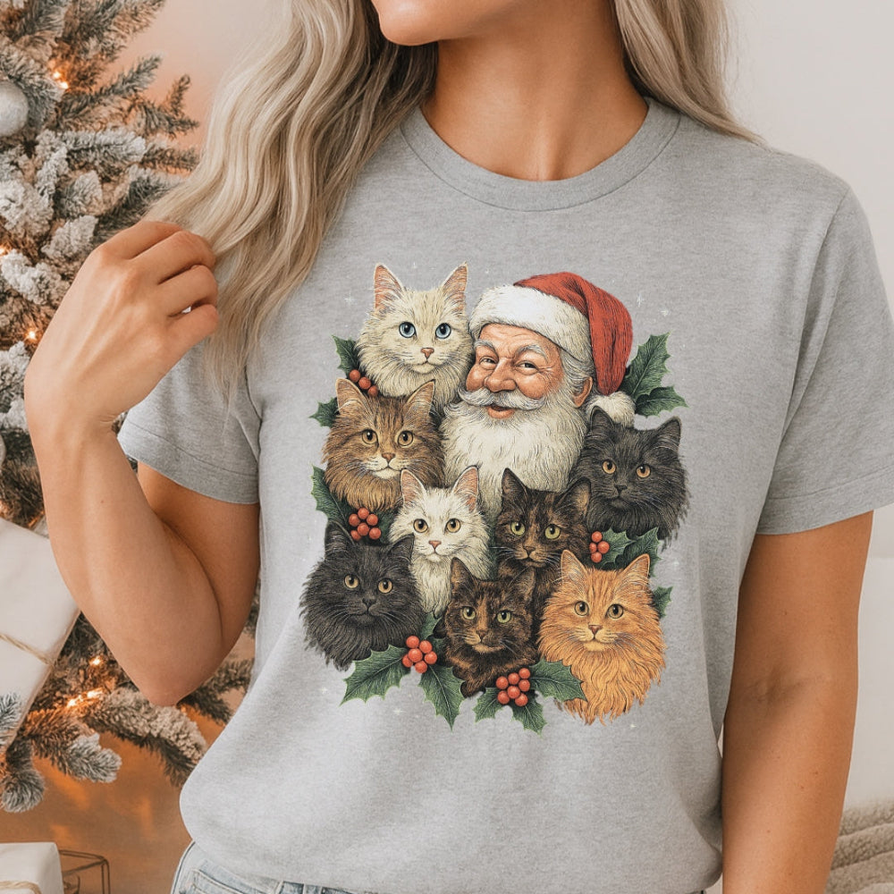 Person wearing a gray t-shirt with a Christmas-themed design of Santa Claus and cats, standing in front of a decorated Christmas tree.