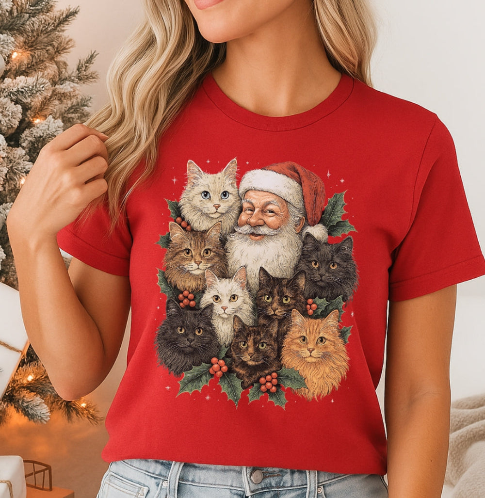 Person wearing a red t-shirt with a Christmas-themed design of Santa Claus and cats, standing in front of a decorated Christmas tree.