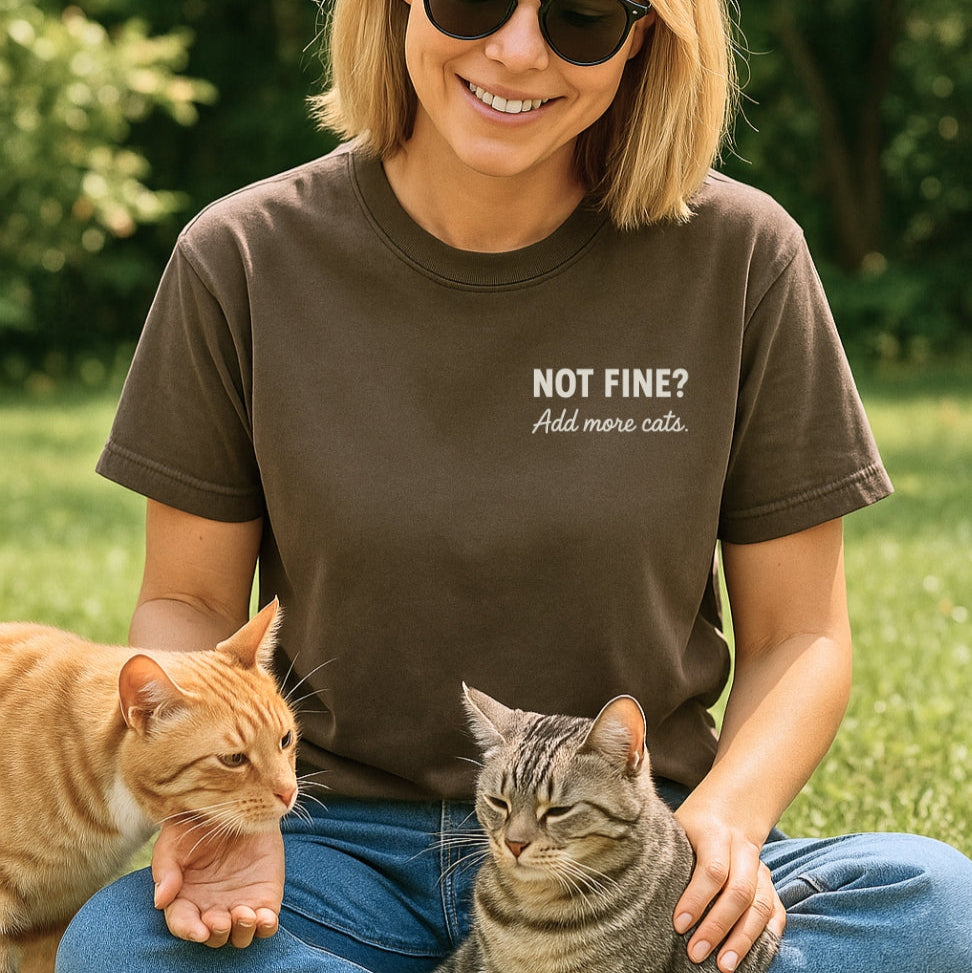 Woman wearing a t-shirt with 'Not Fine? Add more cats' text, sitting outdoors with two cats.