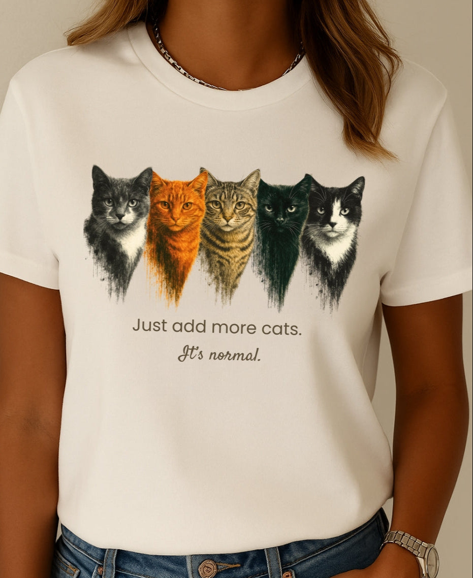 Person wearing a white t-shirt with a graphic of five cats and text 'Just add more cats. It's normal.'