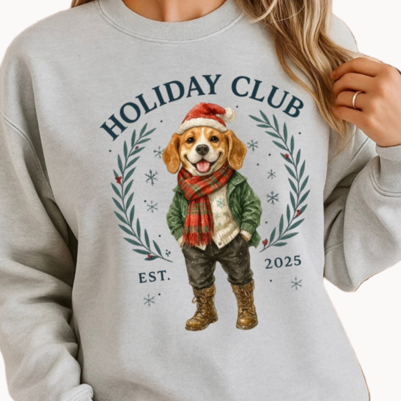 Person wearing a gray sweatshirt with a holiday-themed design featuring a dog and 'Holiday Club' text, standing in front of a decorated Christmas tree.