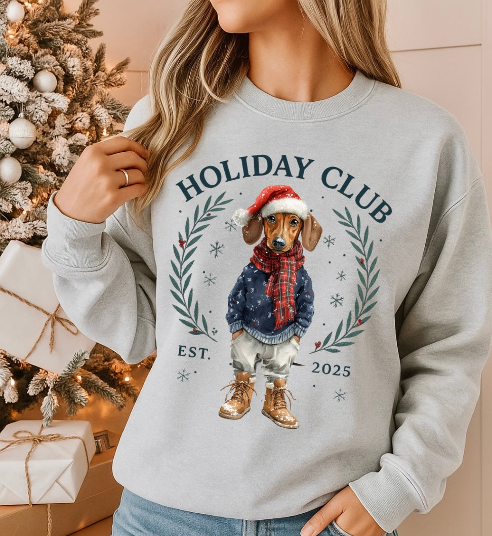 Person wearing a gray sweatshirt with a Christmas-themed design featuring a dog and 'Holiday Club' text, in front of a decorated Christmas tree.