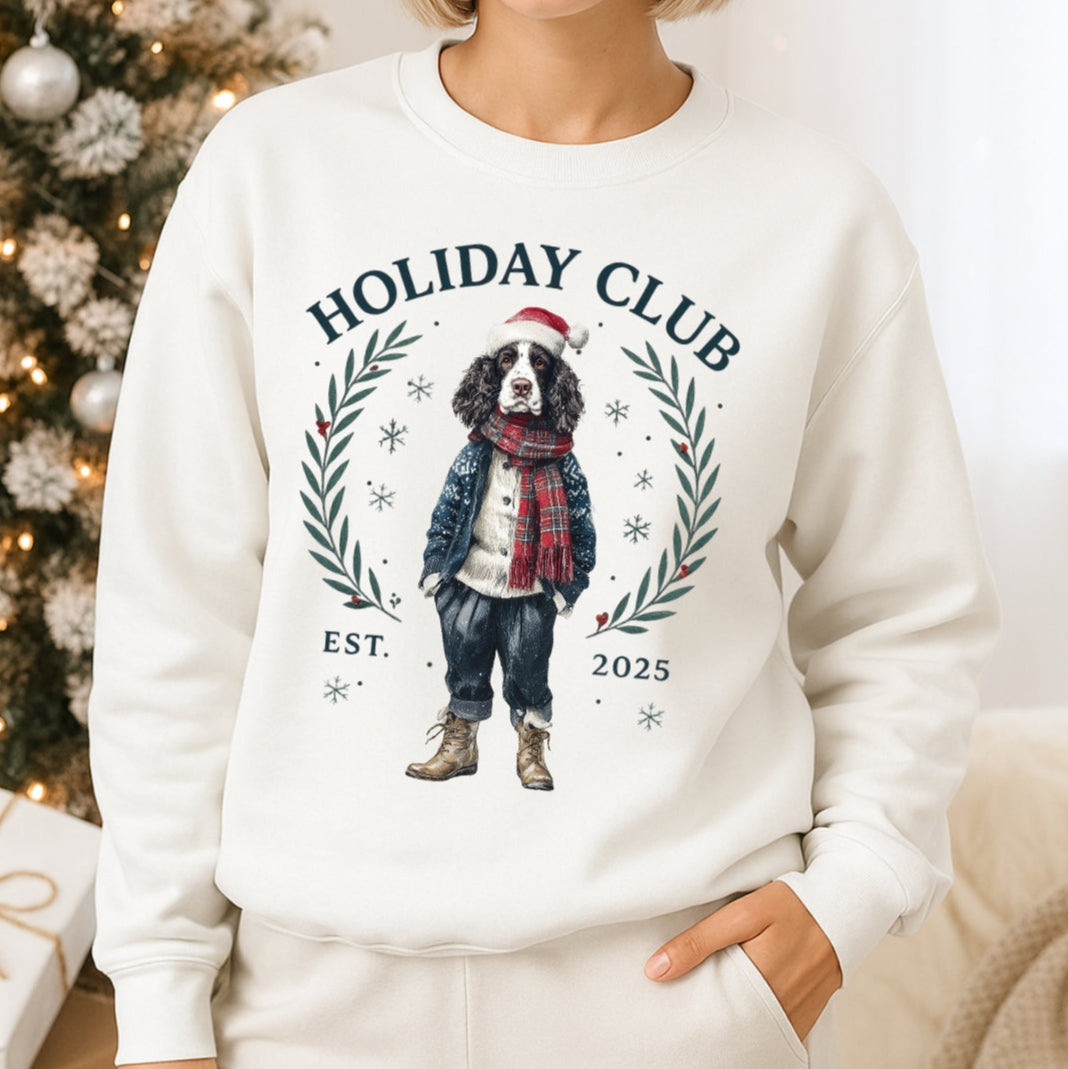 Person wearing a white sweatshirt with 'Holiday Club' and a dog graphic, standing in front of a decorated Christmas tree.