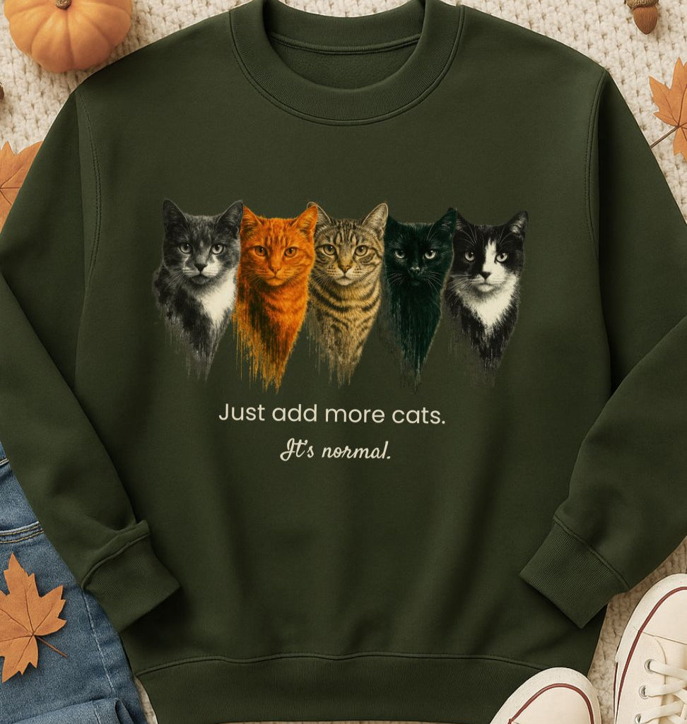 Green sweatshirt with cat illustrations and text 'Just add more cats. It's normal.' on a textured surface with autumn leaves.
