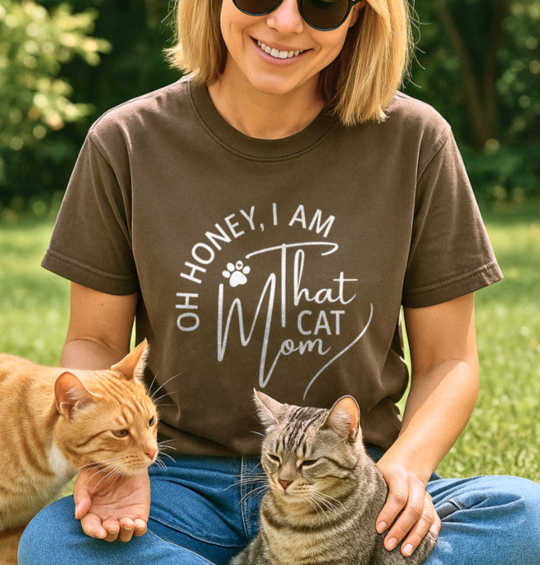 Woman wearing a t-shirt with a cat-themed message, sitting outdoors with two cats.