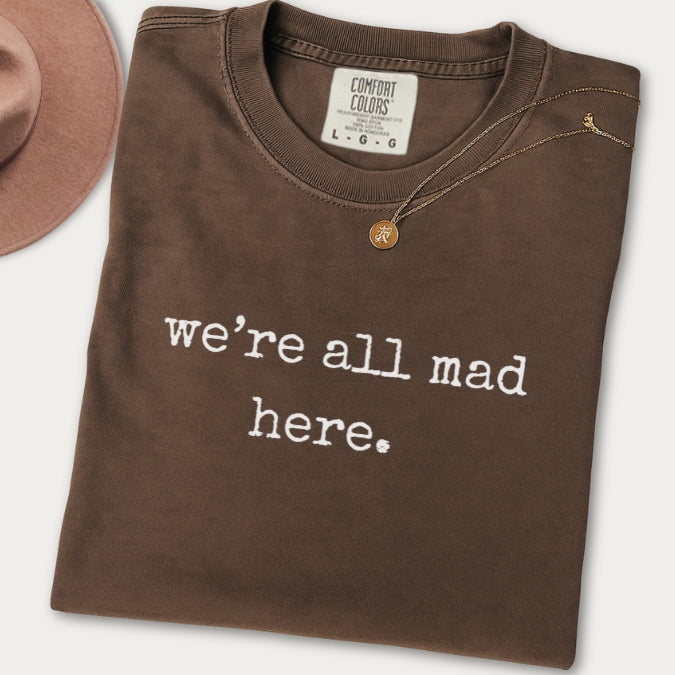 Folded brown sweatshirt with 'we're all mad here' text on a white background