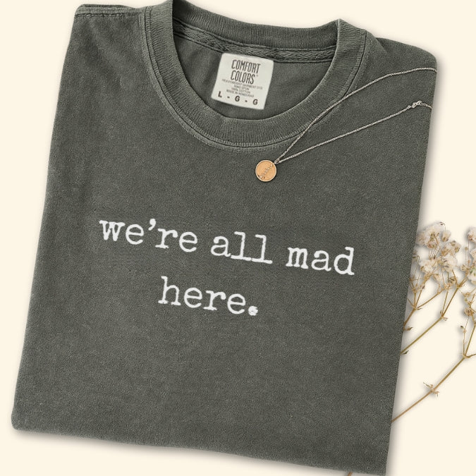 Folded dark gray t-shirt with 'we're all mad here' text on a light background