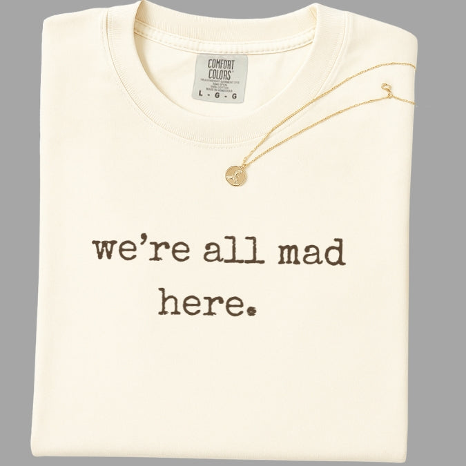 Folded cream-colored shirt with 'we're all mad here.' text and a gold necklace on a gray background