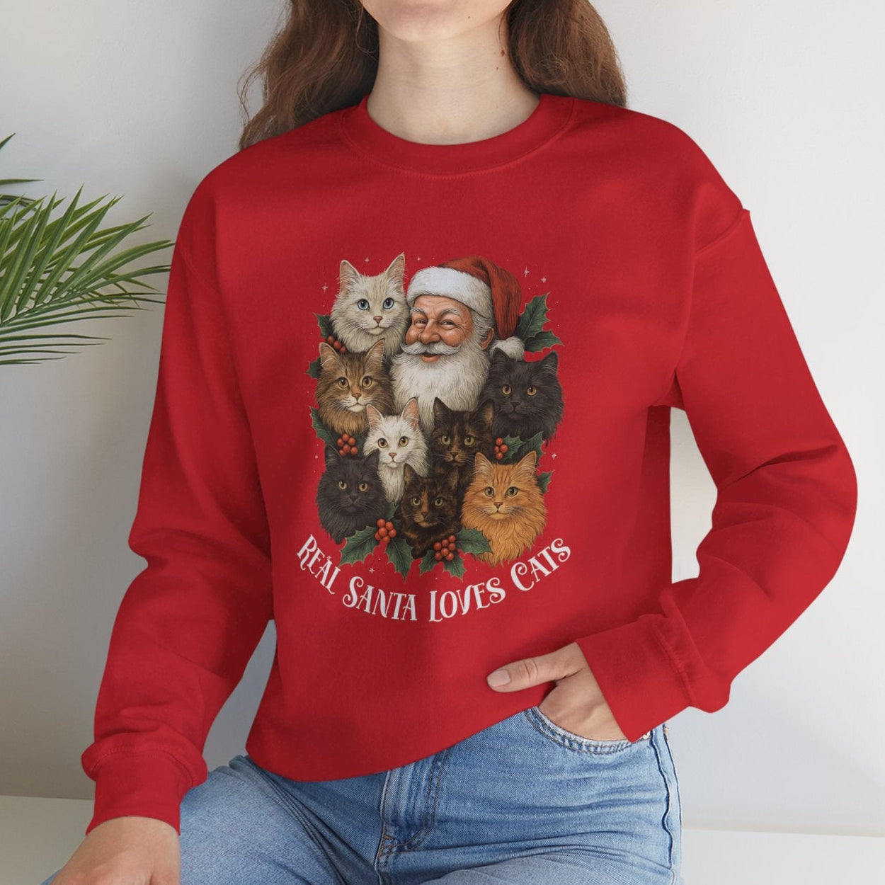 Person wearing a red Christmas-themed sweatshirt with Santa and cats, sitting on a white surface with a plant in the background.
