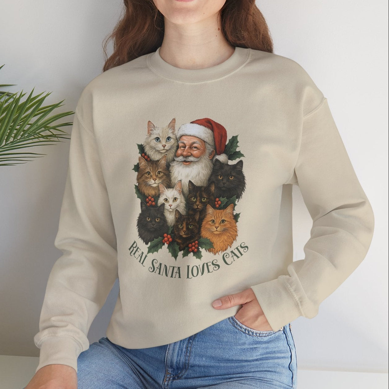 Woman wearing a beige sweater with a graphic design of Santa Claus and cats, sitting on a white surface with a plant in the background.