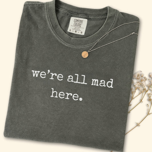 Folded dark gray t-shirt with 'we're all mad here' text on a light background