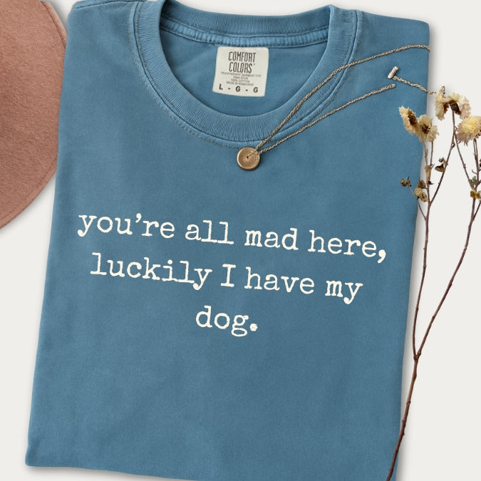 Blue t-shirt with text 'you're all mad here, luckily I have my dog.' on a white background.
