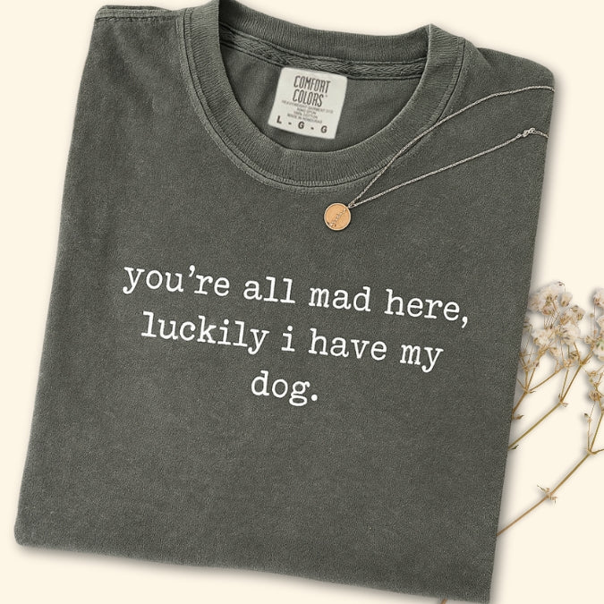 Folded dark Comfort Colors pepper gray t-shirt with text 'you're all mad here, luckily i have my dog.' on a light background.